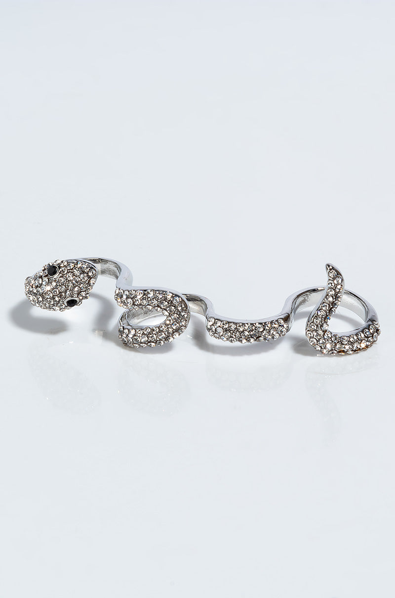 The IN THE GRASS SNAKE RHINESTONE STATEMENT RING, designed to wrap around two fingers in the shape of a silver snake, is adorned with sparkling stones that give it a glittery appearance. The head of the snake features dark gemstones for eyes. The ring is showcased against a plain, light background.