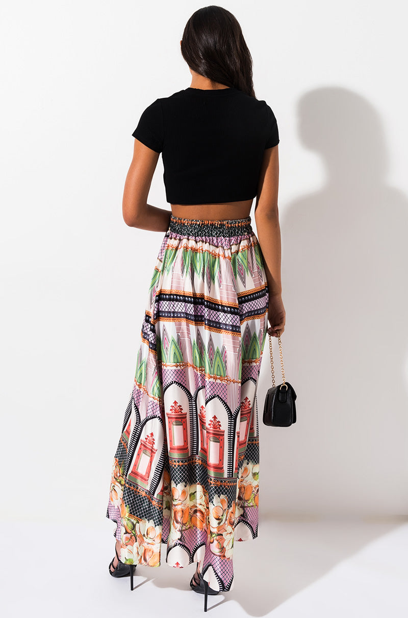 A woman stands facing away from the camera, wearing a black crop top and Caribbean Queen, Inc.'s SEXY MAXI SKIRT—a long, colorful skirt adorned with intricate patterns and images like floral motifs and perfume bottles. She carries a small black purse and wears high-heeled sandals.