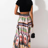 A woman stands facing away from the camera, wearing a black crop top and Caribbean Queen, Inc.'s SEXY MAXI SKIRT—a long, colorful skirt adorned with intricate patterns and images like floral motifs and perfume bottles. She carries a small black purse and wears high-heeled sandals.