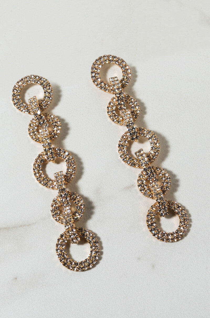 The ISLAND RHINESTONE CHAIN EARRING is a pair of dangling earrings that showcases a series of linked circular hoops, each adorned with sparkling crystals. Each earring features five interconnected hoops, presented against a light, neutral backdrop.