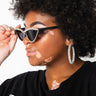 A person with vitiligo wearing ITALY RHINESTONE FRAME SUNNIES and large hoop earrings. They have curly hair and are wearing a black shirt with white text. The person is adjusting their sunglasses while looking to the side.