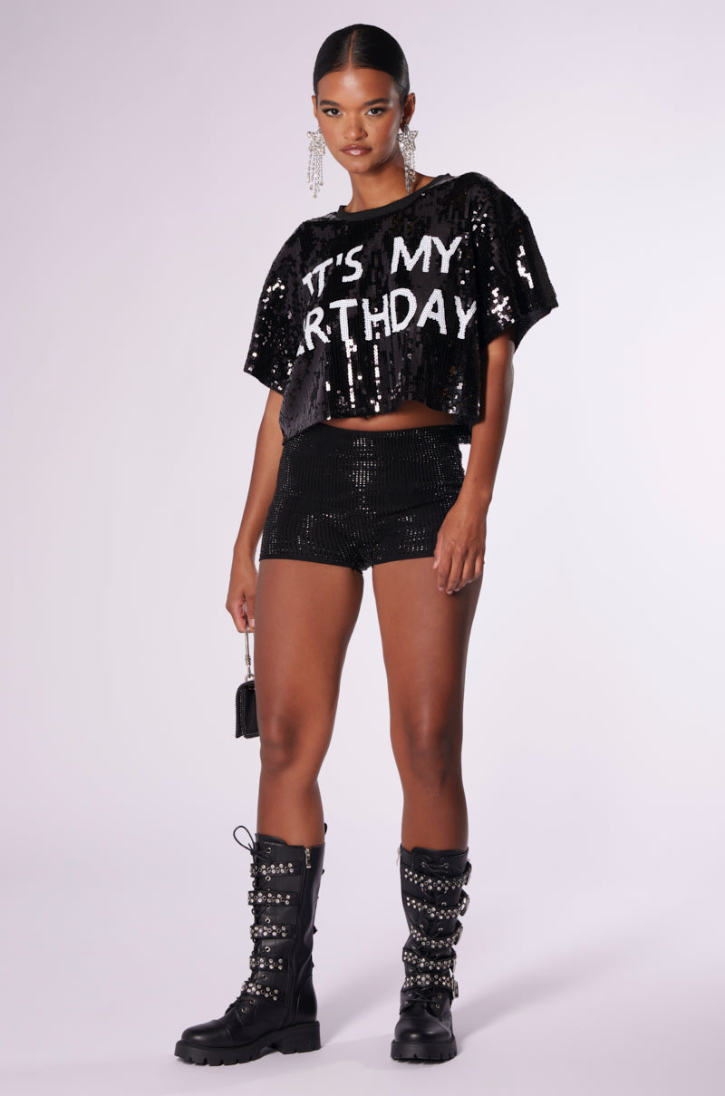 A person wearing an "IT'S MY BIRTHDAY" sequin T-shirt, paired with knee-high lace-up boots and long, sparkling earrings, holds a small black purse while standing against a plain background.