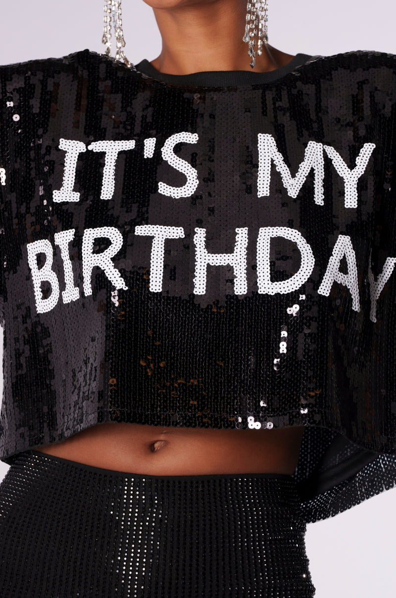 Person wearing an IT'S MY BIRTHDAY SEQUIN T-SHIRT in black, featuring white sequins and a cropped silhouette. The top is paired with matching black sequined pants. Silver dangling earrings are partially visible.
