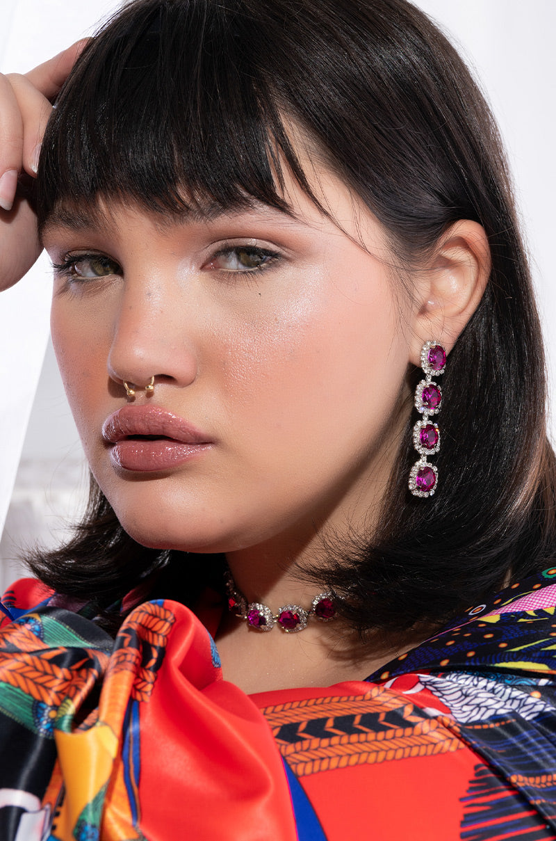A person with medium-length dark hair and bangs wears IT'S THE PINK EARRINGS FOR ME, a matching necklace, and a gold septum ring. They are dressed in a colorful, patterned garment and are posing with a serious expression against a light background.