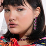 A person with medium-length dark hair and bangs wears IT'S THE PINK EARRINGS FOR ME, a matching necklace, and a gold septum ring. They are dressed in a colorful, patterned garment and are posing with a serious expression against a light background.