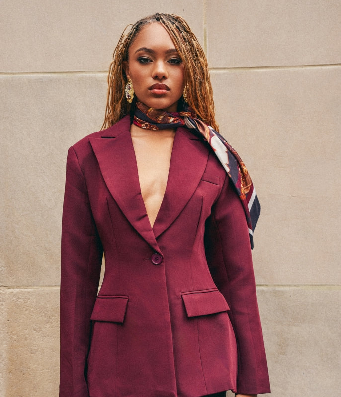 A woman wearing a structured burgundy blazer with a deep V-neckline, paired with a patterned scarf, standing against a beige stone wall.