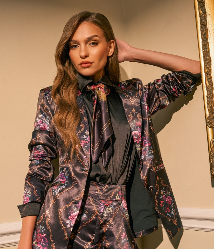 A woman models a matching floral satin blazer and trousers set with a black satin bow tie, posing against a beige wall next to a gold-framed mirror.