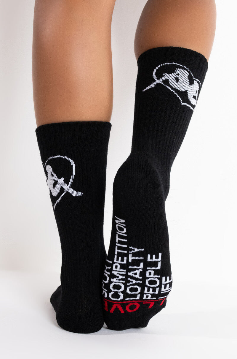A person is wearing the Kappa Endless Love Ankle Socks, which are black with white graphics and text. One sock features a white logo depicting a face in a hat, while the other showcases the words "FOCUS," "COMPETITION," "LOYALTY," "PEOPLE," and "LIFE" in white lettering. 0