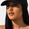 A person with long, dark hair wearing the SEXY TREND HAT, featuring a black visor with a white Kappa logo, gazes into the distance. The plain white background accentuates their features and highlights the hat.