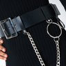 A close-up image of a person wearing a black ribbed sweater, highlighting the KEEP THE CHAIN-GE SINGLE CHAIN BELT. The wide black leather belt features a large silver buckle, adorned with a silver chain and metal ring accessories hanging from it. Part of the person's hand is visible resting on their hip.