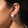 Close-up of a person's ear adorned with the elegant KEIRA COIN EARRING. The earring features a large, smooth pearl at the top with a detailed gold disc hanging below it, crafted with intricate patterns. The person has dark hair pulled back.