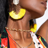 A person wearing a colorful outfit adorned with a tropical print and polka dots is accessorized with the LEO PAVE LAYERED NECKLACE, featuring gold and silver layers, one with the word "Love." They also sport large, dangling earrings with yellow tassels and metallic leaf designs.