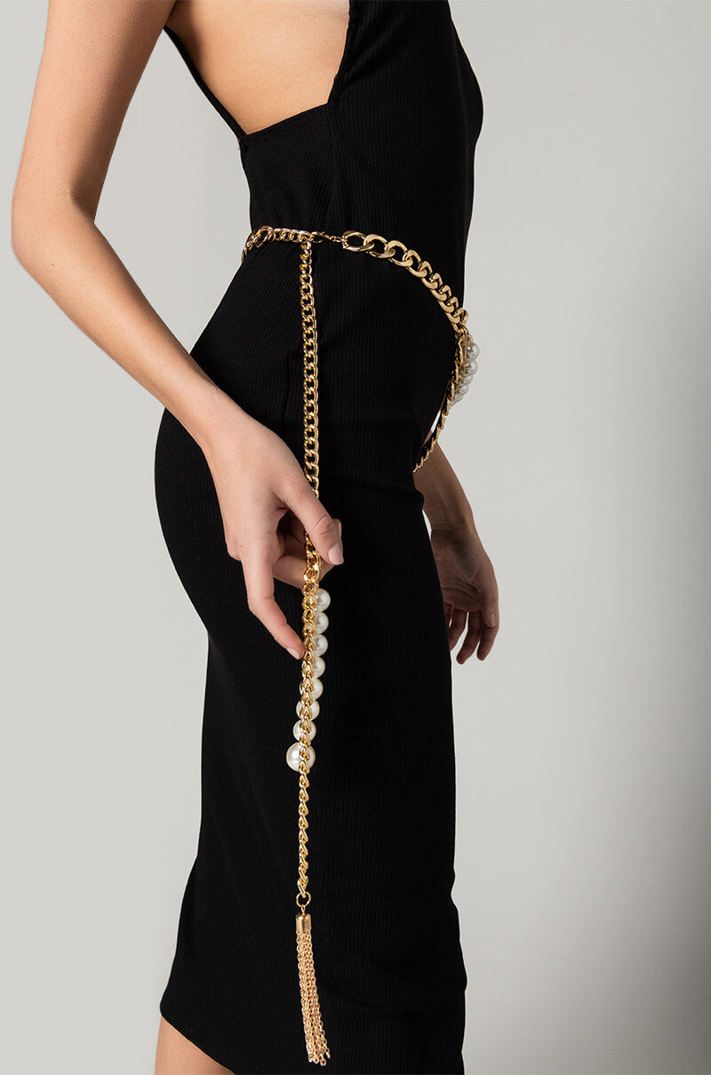 A woman wearing a form-fitting black dress accessorized with the LIFE IS GOLDEN CHAIN PEARL BELT, which features pearl accents and tassels on the end, stands against a plain background. She is posed with one arm slightly bent and resting along her side.