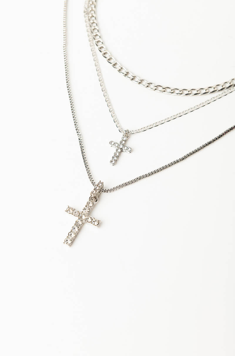 Three silver necklaces from the LIKE A PRAYER LAYERED NECKLACE collection are displayed vertically on a white background. The top two necklaces feature thinner chains, with the uppermost having a simple chain design and the middle one bearing a small cross pendant. The bottom necklace, thicker in design, also has a cross pendant adorned with small crystals.