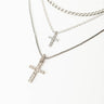 Three silver necklaces from the LIKE A PRAYER LAYERED NECKLACE collection are displayed vertically on a white background. The top two necklaces feature thinner chains, with the uppermost having a simple chain design and the middle one bearing a small cross pendant. The bottom necklace, thicker in design, also has a cross pendant adorned with small crystals.