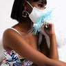 A woman with a short bob haircut wears colorful earrings and a patterned outfit while sitting on a white chair. She sports the LIL BIT EXTRA FASHION FACE COVER, a white face mask adorned with light blue feathers, covering her nose and mouth. The background is a plain white wall.