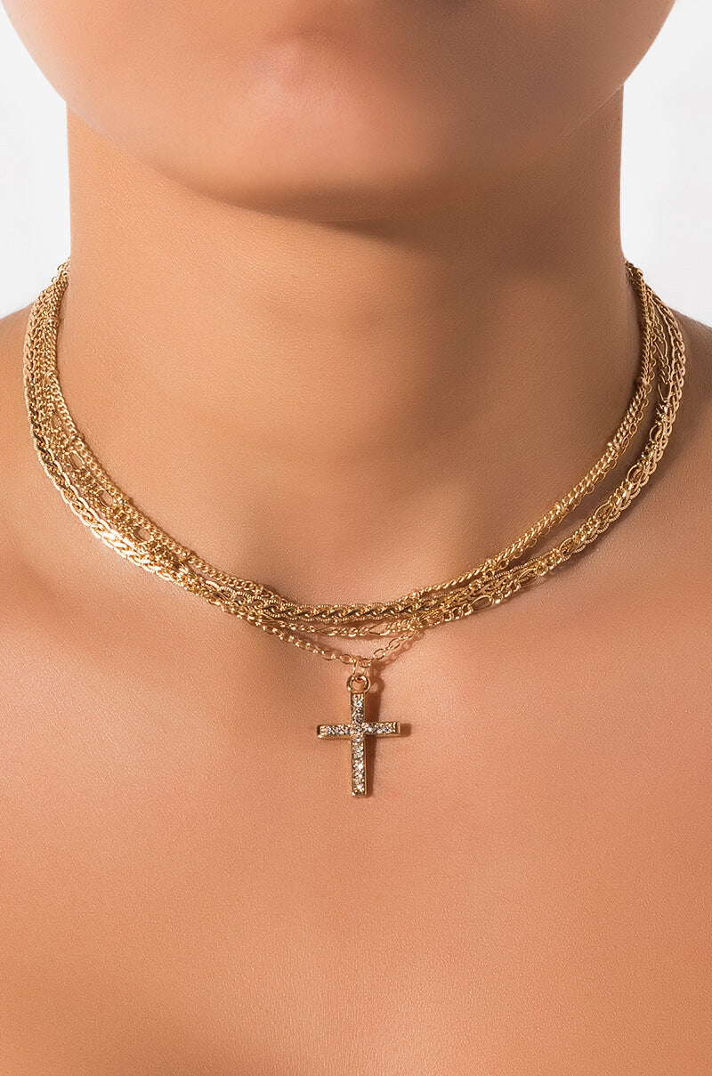 Close-up of a person wearing the LORD KNOWS CHOKER SET, featuring multiple gold chains and a pendant in the shape of a cross embellished with small gemstones. The jewelry exudes elegance and is the focal point of the image against a plain background.