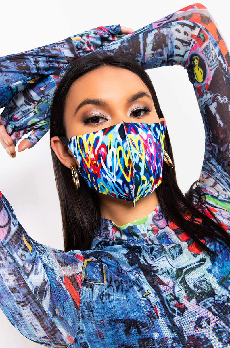 A woman with long dark hair wears the "LOVE ON THE WALL FASHION FACE COVER," featuring a vibrant abstract pattern of hearts and various shapes. She accessorizes with hoop earrings and is dressed in a vivid, patterned, long-sleeved top. Her arms are positioned behind her head, highlighting both her outfit and the artistic design of the face cover.