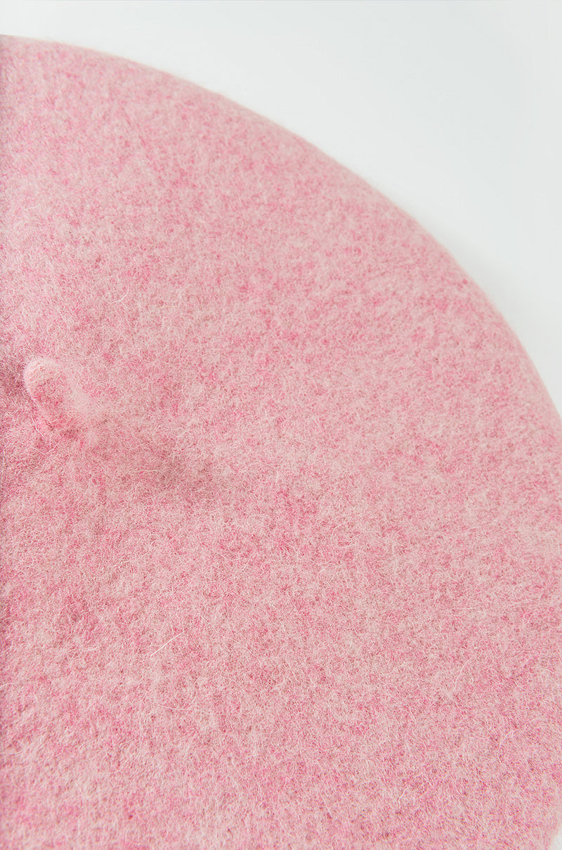 Close-up image of the MACAROON BERET HAT IVORY made from a soft, fuzzy, felt-like material in a pastel pink hue. The beret features a small nub or stalk in the center, characteristic of this style of hat. The background is a plain, light surface.