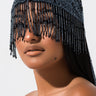 A woman with long dark hair wears the MAKE ME THIS WAY BEADED HAT in gold, which features intricately beaded details and dangling strands that partially cover her eyes and forehead. The plain white background highlights her smooth complexion and the ornate design of the hat.