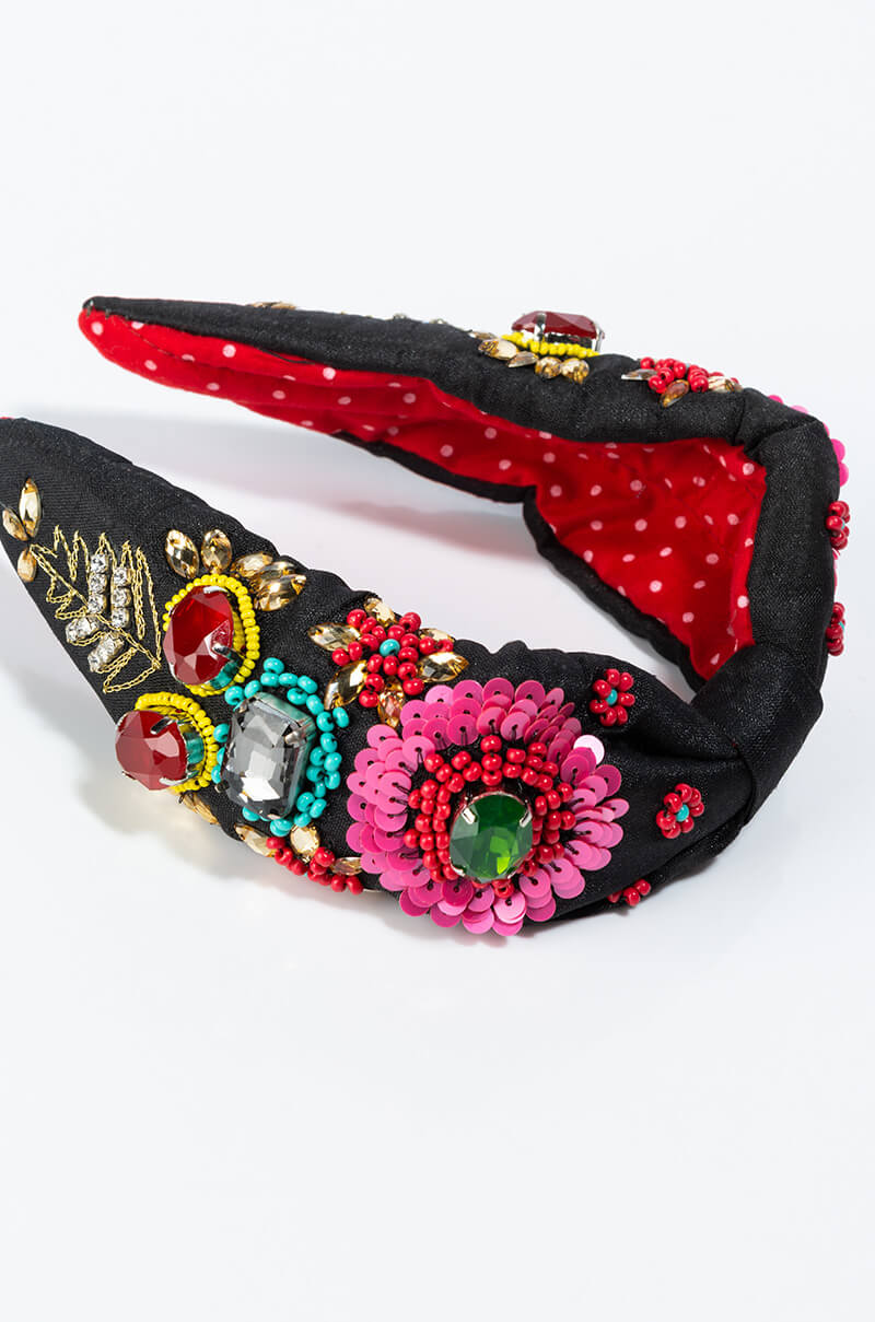 Introducing the MAMACITA EMBELLISHED HEADBAND, a black padded headband adorned with vibrant embroidery, sequins, and an array of gemstones in shades of red, green, blue, and gold. The inner lining features a cheerful red fabric with white polka dots. This stunning accessory showcases intricate floral and leaf motifs throughout its design.