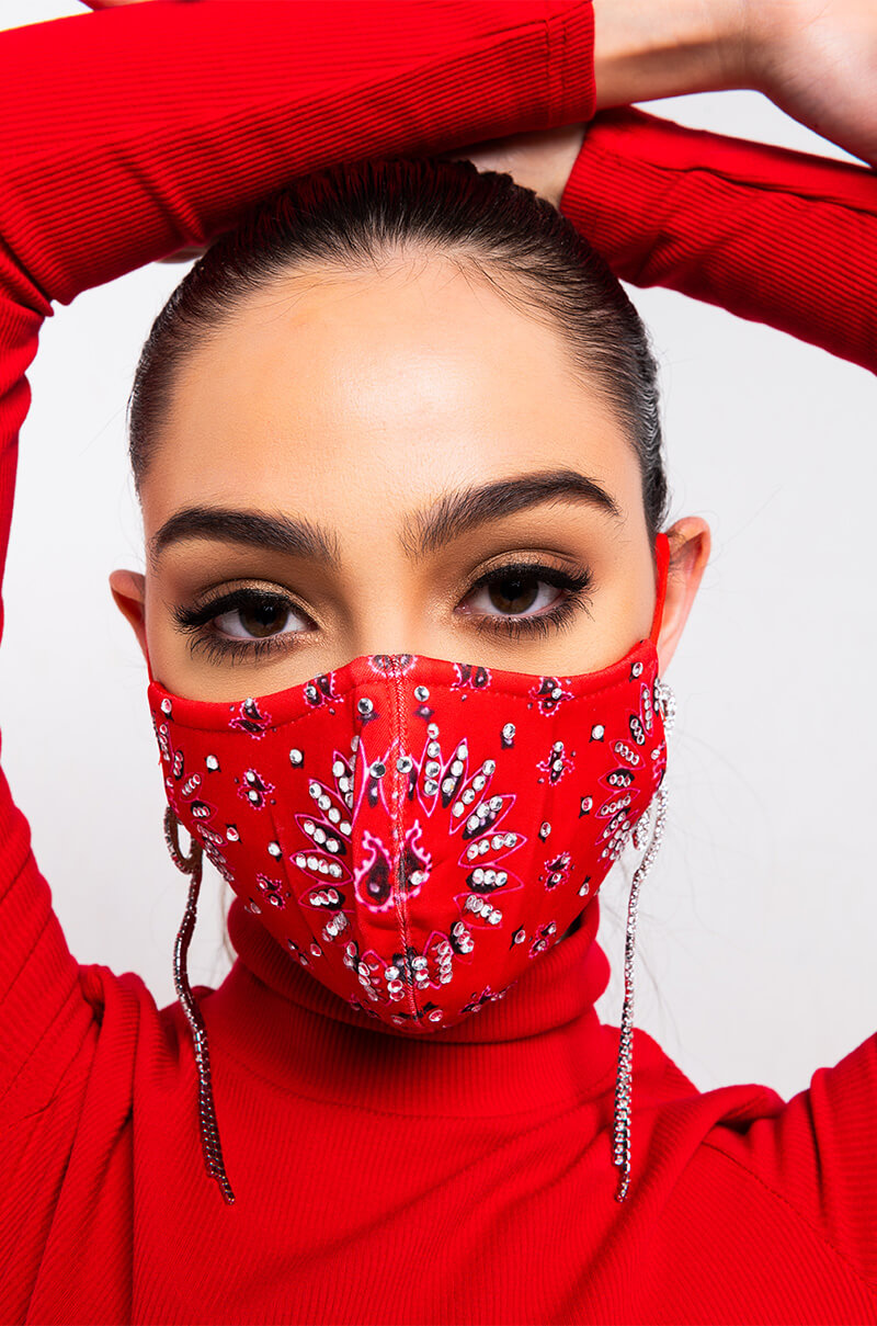 A person wearing the MATERIALGIRL RHINESTONED FASHION FACE COVER in red and a matching red turtleneck, posing with their arms raised over their head. The face cover is adorned with intricate jeweled patterns. The person also has neatly styled eyebrows and long, dangling earrings.