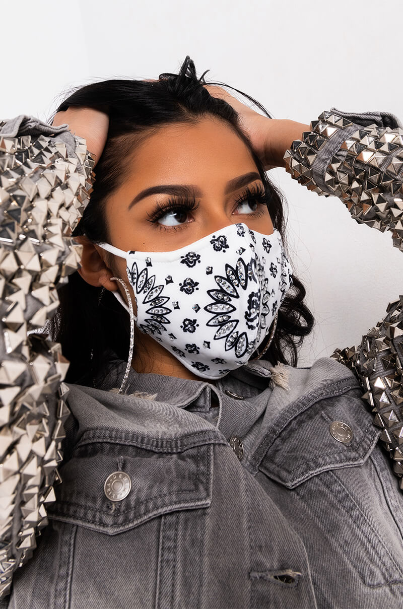 A person is seen wearing the MATERIALGIRL RHINESTONED FASHION FACE COVER, which features a white and black pattern. They are also sporting a jacket adorned with silver spikes on the sleeves. With dark hair and their hands resting on their head, they pose against a plain, light-colored background.