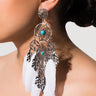 Close-up of a person wearing the MIKAYLA DREAMCATCH EARRING, designed to resemble a dreamcatcher. The earring showcases small turquoise beads and multiple dangling elements, including silver leaves and white feathers. The person's hair is pulled back, set against a plain white background.