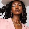 A woman with long, wavy black hair wears a wide-brimmed white hat, a pink blazer, and the RILEY PAVE CHAIN NECKLACE GOLD from Jennifer Accessories. She accessorizes with vibrant pink and gold drop earrings. The background is light gray as she looks confidently at the camera.