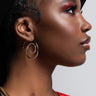 Profile view of a person showcasing the MOVE YOUR BODY LIKE A SNAKE MA RHINESTONE SNAKE HOOP with a black wide-brimmed hat, large hoop earrings, and a gold necklace. The person has a smooth complexion, eye makeup with bold red eyeshadow, and a serious expression.