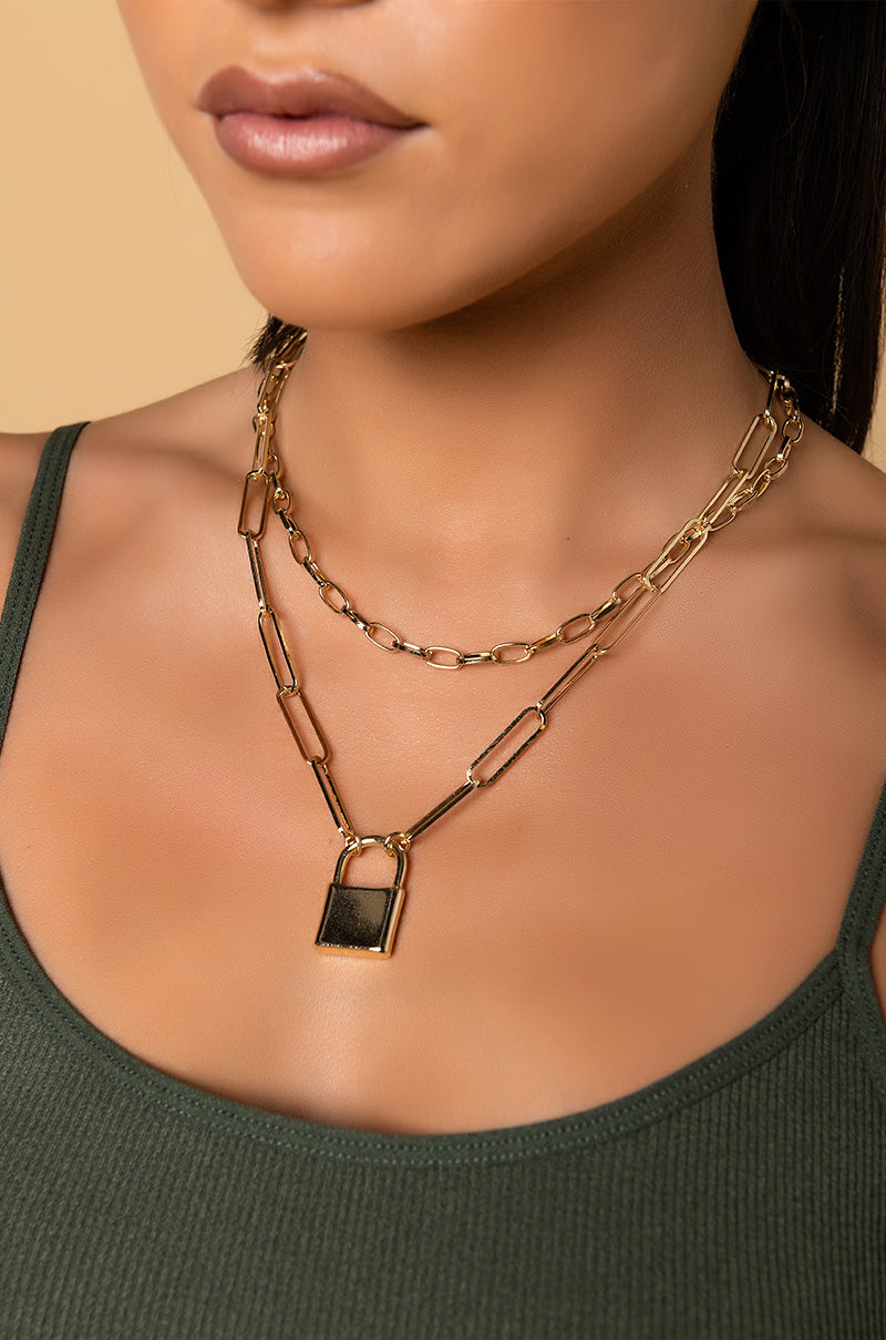 A close-up of a person wearing a sleeveless green top. They are adorned with the NECKLACE GOLD, featuring two layers – one shorter and delicate, and the other with larger links, showcasing a square black pendant. The background is a solid light peach color.