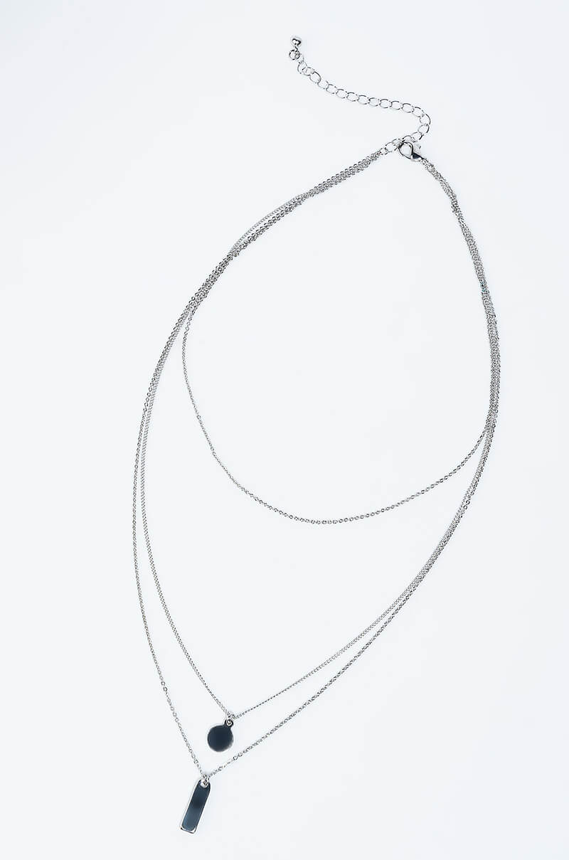 The NECKLACE SILVER, a three-layered silver chain necklace, is elegantly showcased against a white background. The design includes a small black circular pendant, a rectangular black pendant, and a plain chain as the shortest layer. An adjustable chain clasp can be seen at the top.