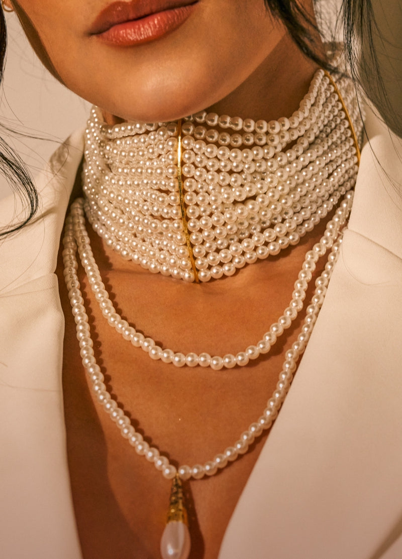 A close-up of a model wearing a multi-strand pearl necklace with gold-toned metal accents and a teardrop pearl pendant, styled with a white blazer.