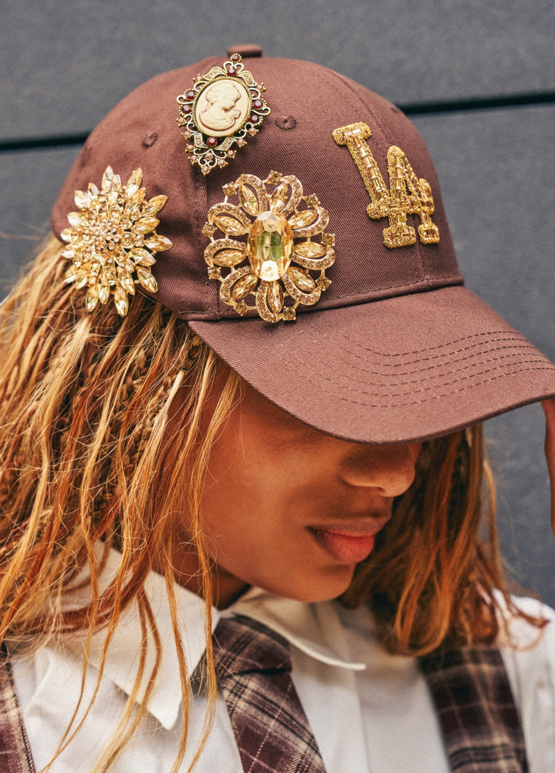 A close-up of a person wearing a brown baseball cap adorned with multiple ornate gold brooches and a gold LA logo patch, paired with a white collared shirt and plaid tie against a dark gray background.