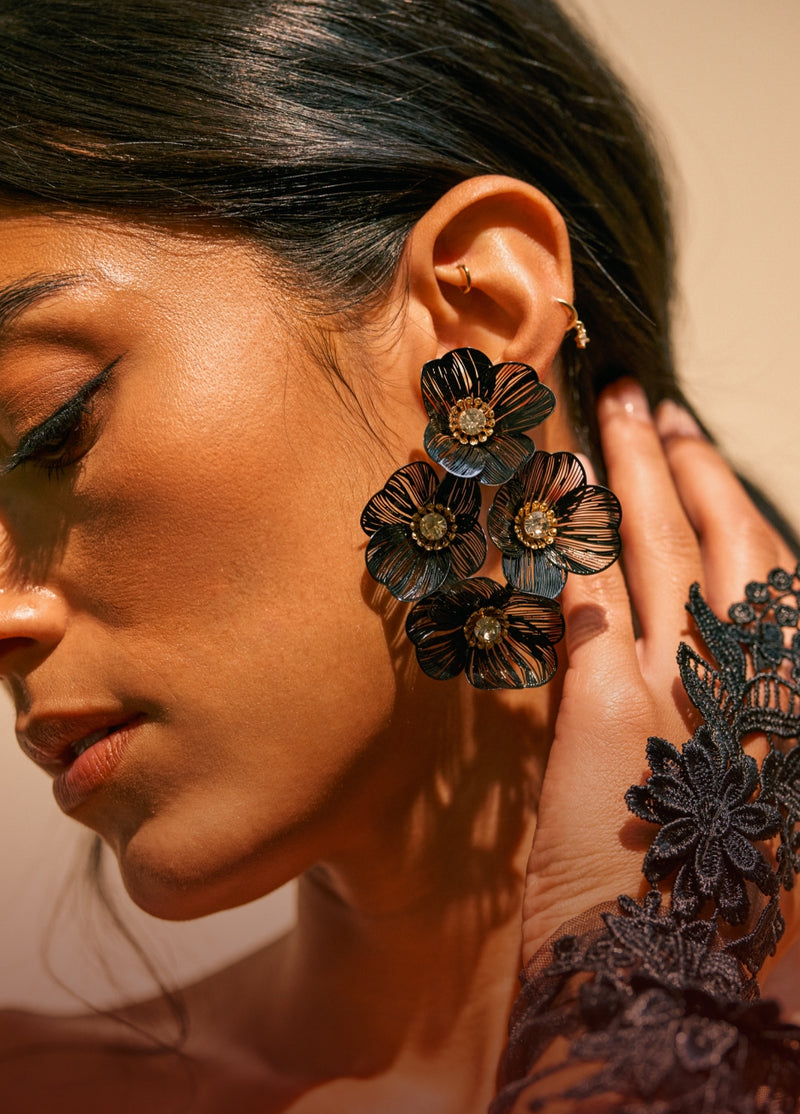 A close-up of a woman wearing large black floral statement earrings with gold-toned center details and delicate gold hoop earrings, against a warm beige background.