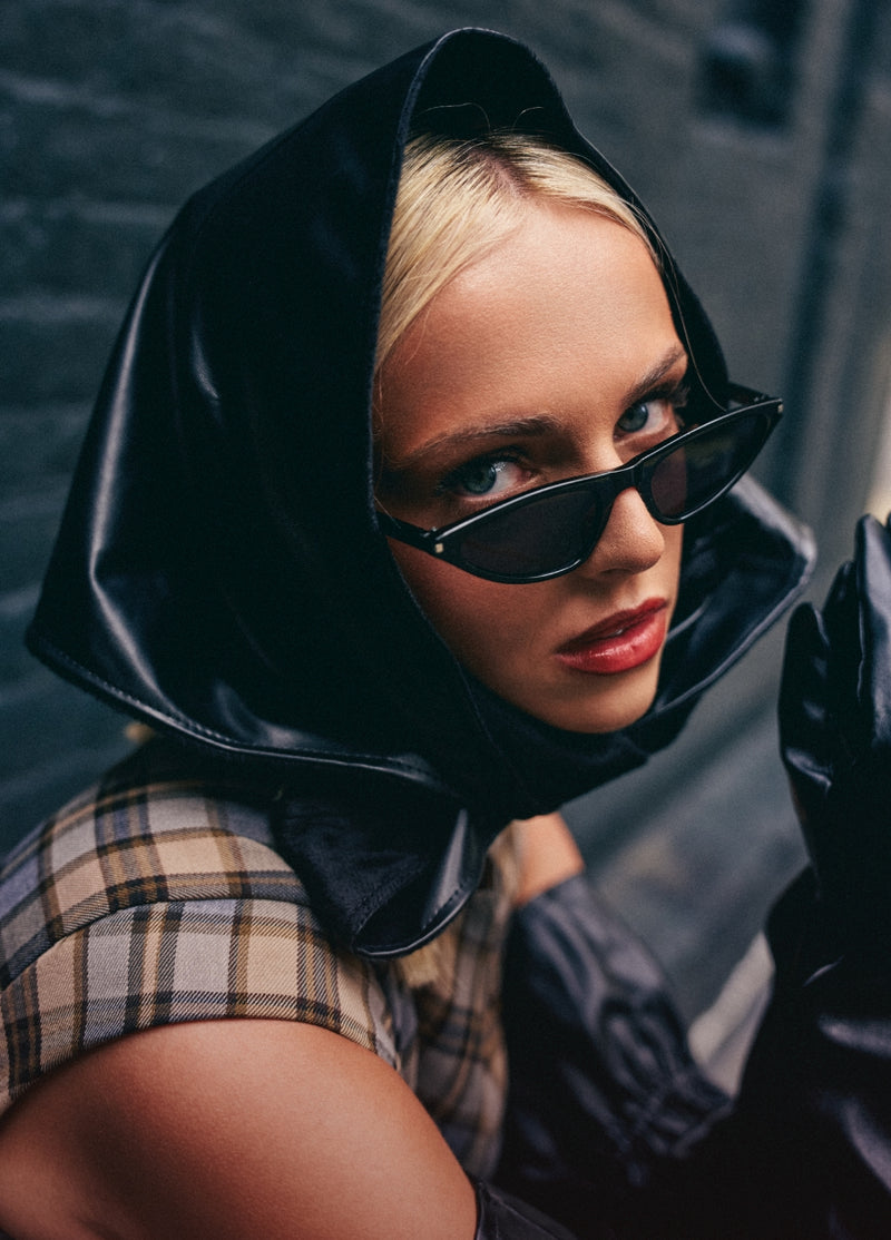A close-up lifestyle shot of a model wearing a black hooded jacket, black sunglasses, and a beige plaid top against a dark gray brick wall.