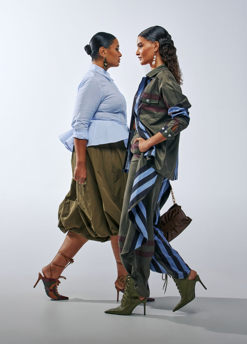 Two models walking side-by-side in a studio setting, showcasing fashion outfits. The model on the left wears a light blue long-sleeve shirt, olive green pleated skirt, and brown strappy heels.