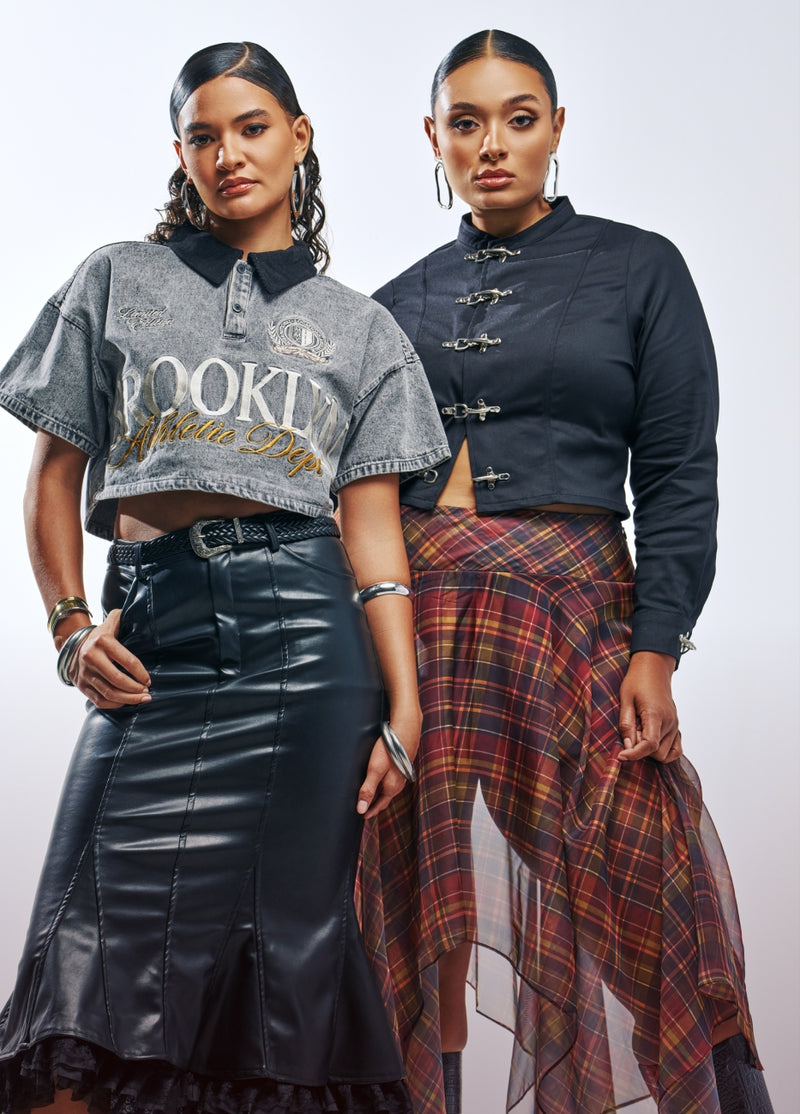 Two models posing in streetwear fashion against a light gray studio background. The model on the left wears a gray graphic crop top with a black collar, a black faux-leather skirt, and silver jewelry.