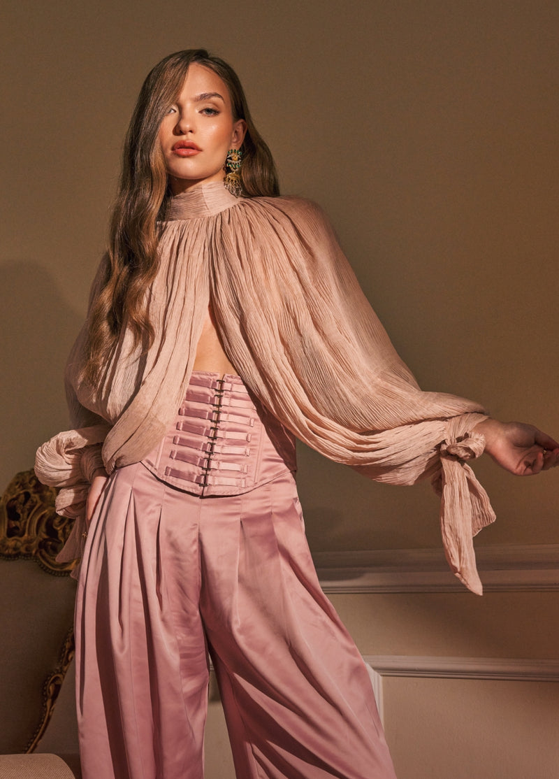 A woman models a dusty pink two-piece outfit consisting of a sheer, high-neck long-sleeve top with dramatic gathered sleeves and matching satin wide-leg trousers, styled with a structured corset-style waist panel.