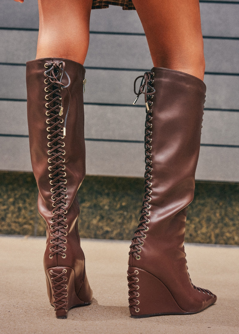 A pair of knee-high brown leather boots with wedge heels and extensive side lacing, shown on a model from the back against a gray paneled wall.
