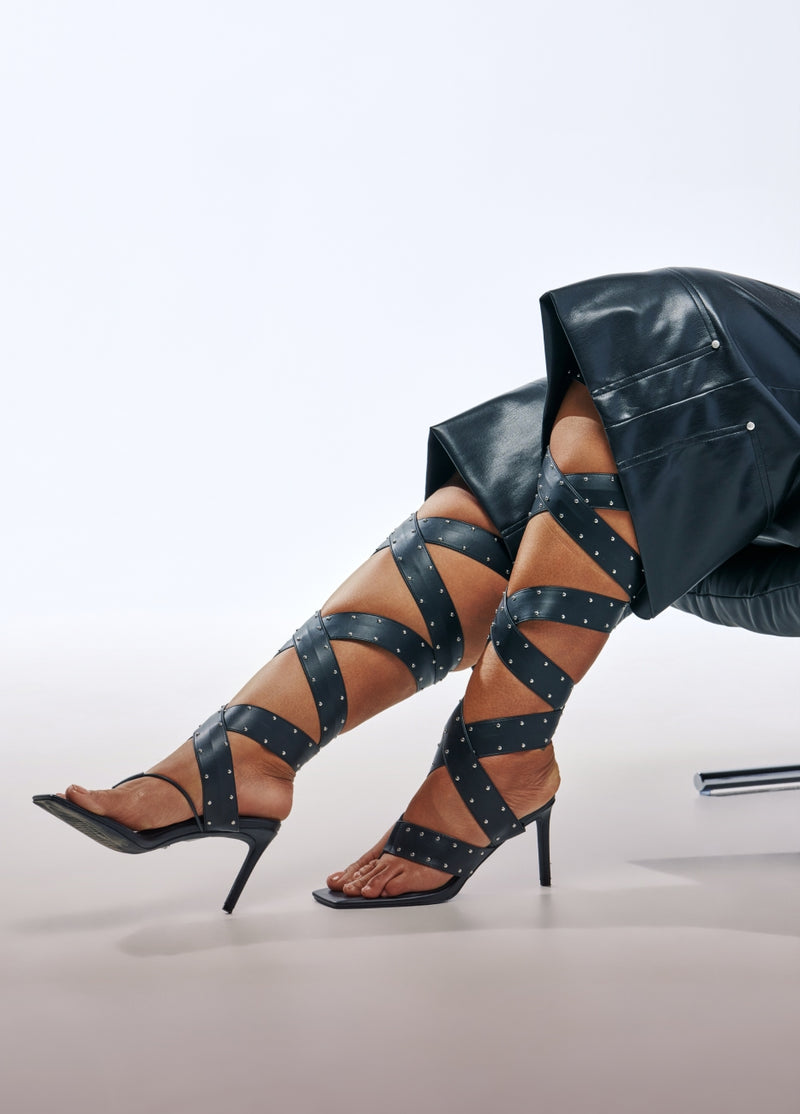 A model's legs wearing black strappy high-heeled sandals with silver studs, paired with a black faux-leather skirt, set against a clean white studio background.