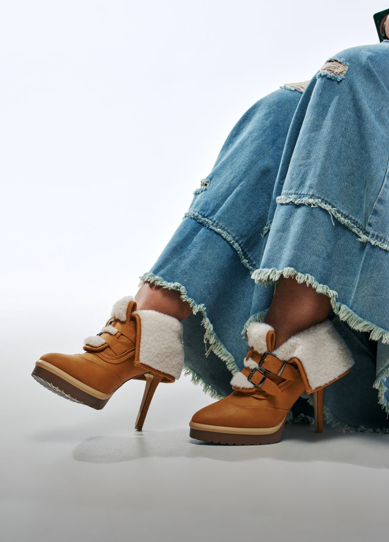 A pair of tan suede high-heeled ankle boots with white shearling lining and double buckle straps, shown on a model wearing distressed light blue jeans against a white studio background.