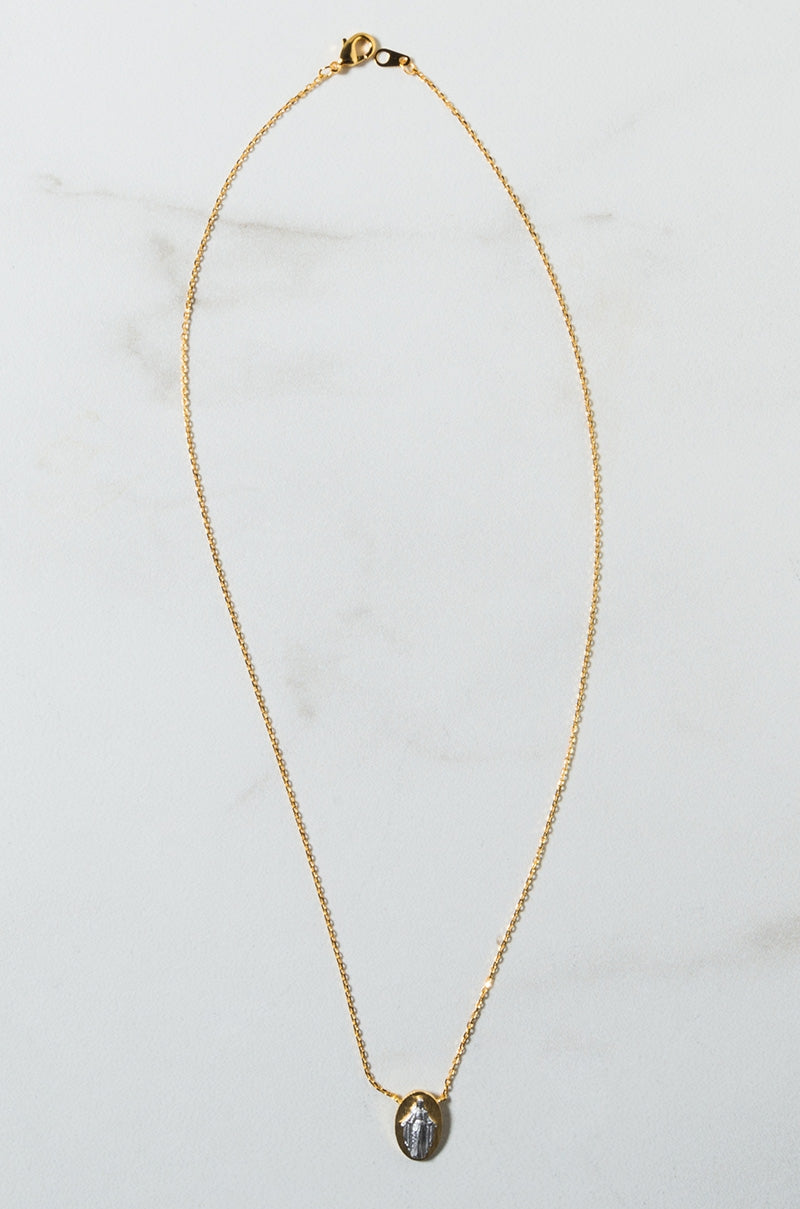 The MARIA DAINTY NECKLACE features a delicate gold chain with a small oval pendant at the center, showcasing a marble-like design. The necklace is elegantly laid flat on a white marble background.