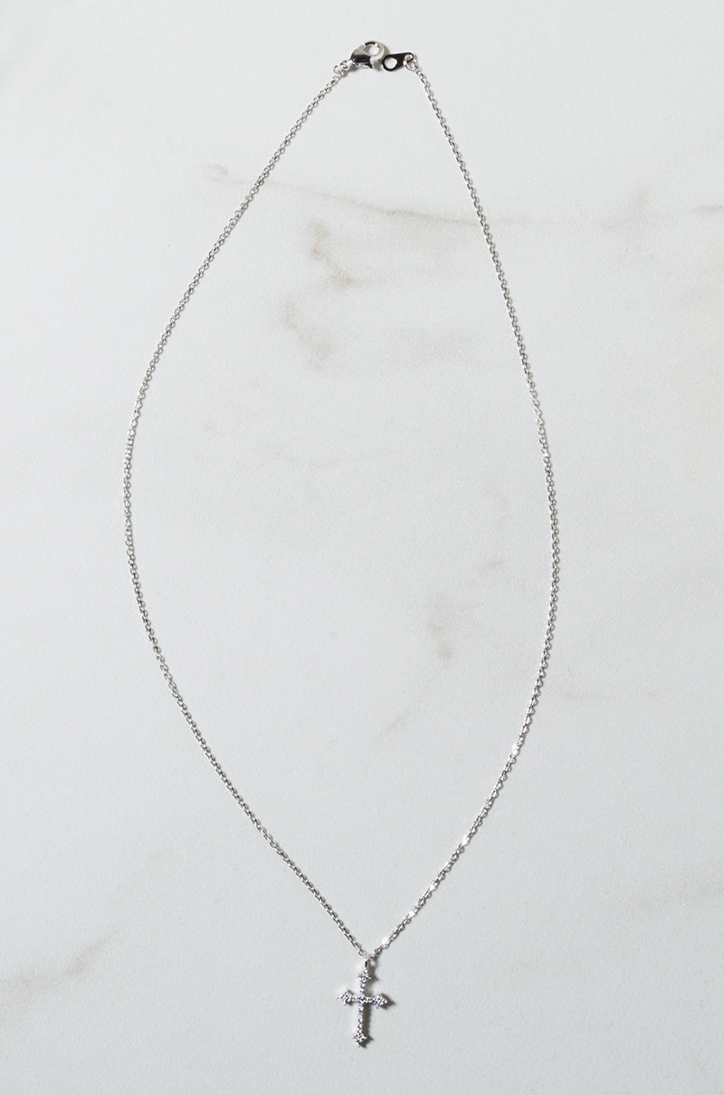 The BLESSINGS NECKLACE, a delicate silver piece featuring a small crucifix pendant, lies on a white background. The necklace boasts a thin and minimalist design, with the cross pendant subtly engraved. The clasp at the top of the chain is also visible.