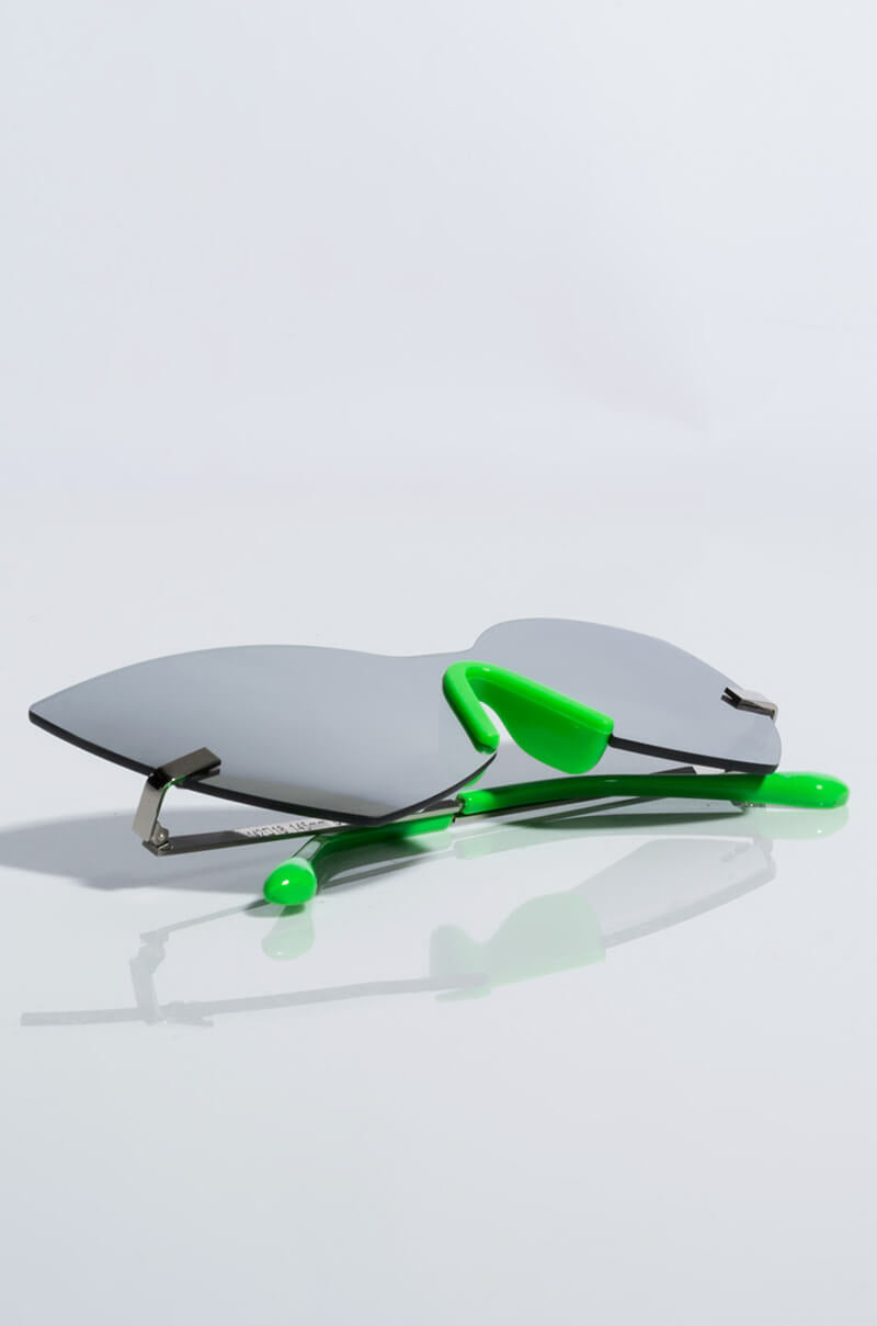 The NO BULLSHIT CAT EYE SUNNIES, featuring sleek reflective silver lenses and vibrant green frames with matching earpieces, lay against a plain white background. Their minimalist design imparts a modern and stylish appearance.