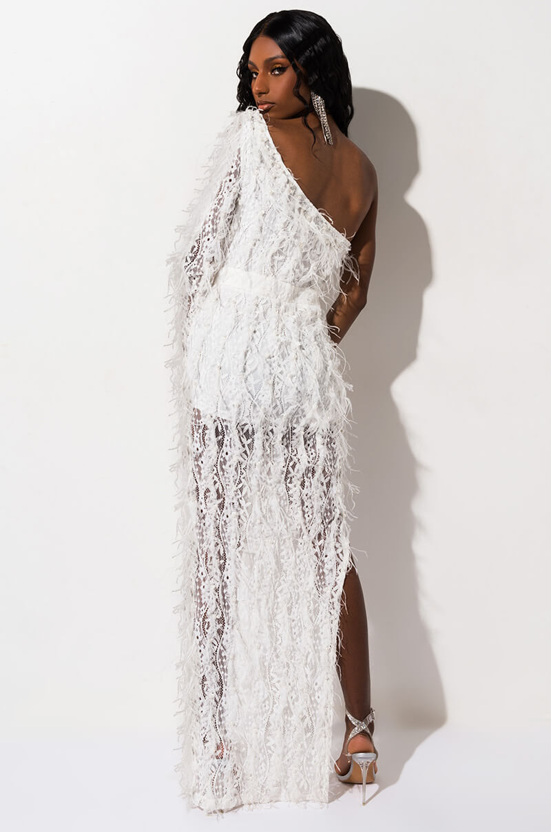 A model stands against a plain white background, facing partially away from the camera. She wears BCCD42084, a white, one-shoulder, semi-sheer dress adorned with textured lace and fringe, and high-heeled silver sandals. Her long black hair cascades down her back.