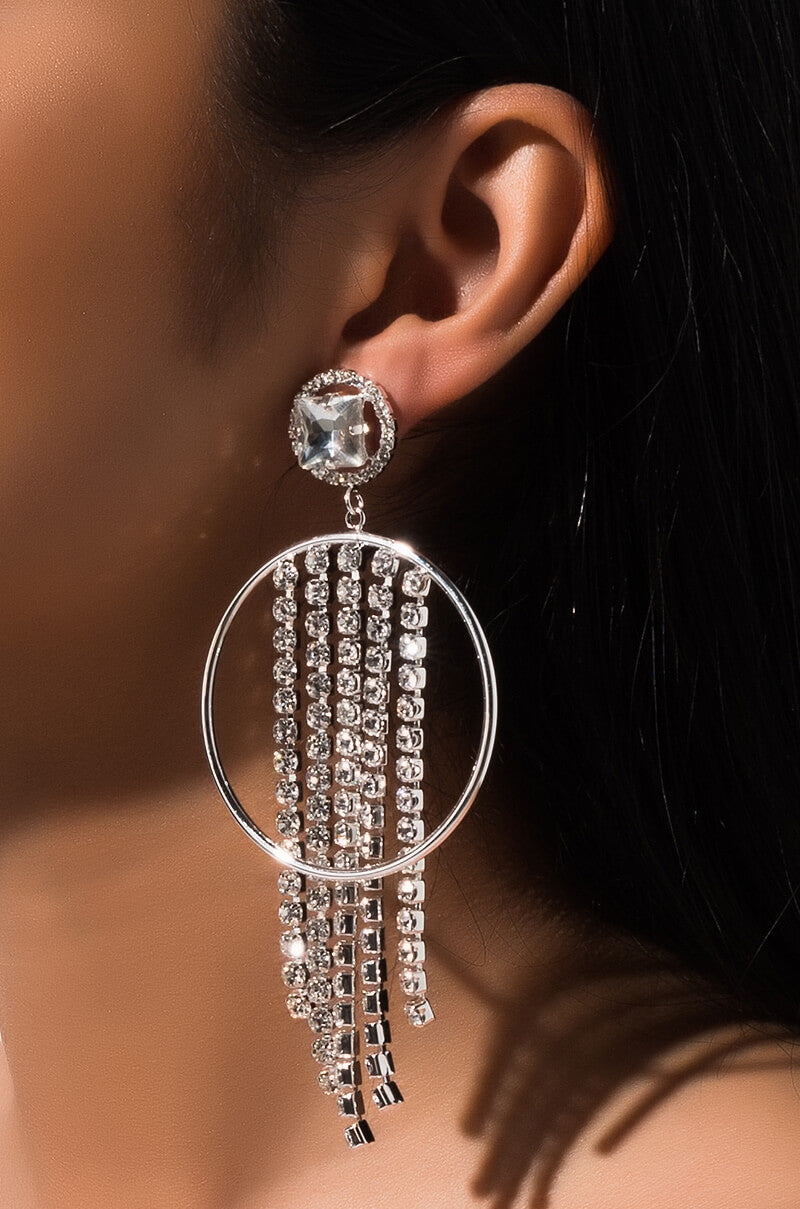Close-up image of a person's ear wearing the SEXY DANGLE EARRING by RM Manufacturing, featuring a large, circular, silver design with multiple dangling strands adorned with small, sparkling crystals. The earring stud showcases a larger, faceted crystal, enhancing the overall glamorous aesthetic.