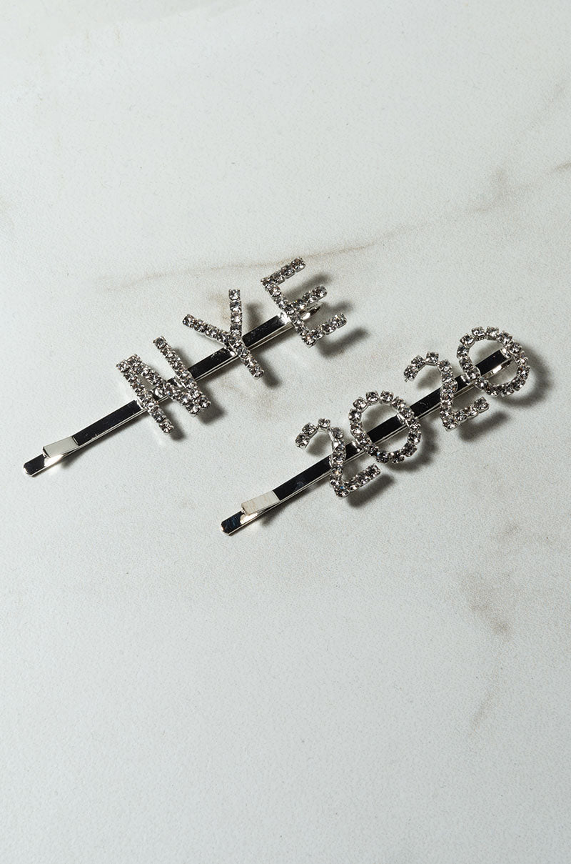 Two metallic SEXY CLIP HAIR accessories lying on a marble surface. One hairpin has the letters "NYE" adorned with small rhinestones, and the other hairpin displays the numbers "2020," also embellished with rhinestones.