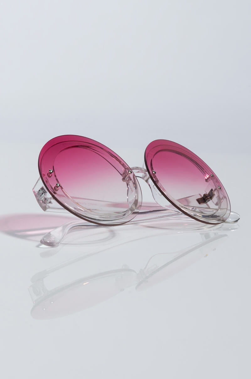 The OMBRE SUNSET ROUND SUNGLASSES, featuring gradient pink lenses and transparent frames, rest on a reflective surface. The lenses gracefully shift from a deep pink at the top to clear at the bottom, casting a delicate shadow below.