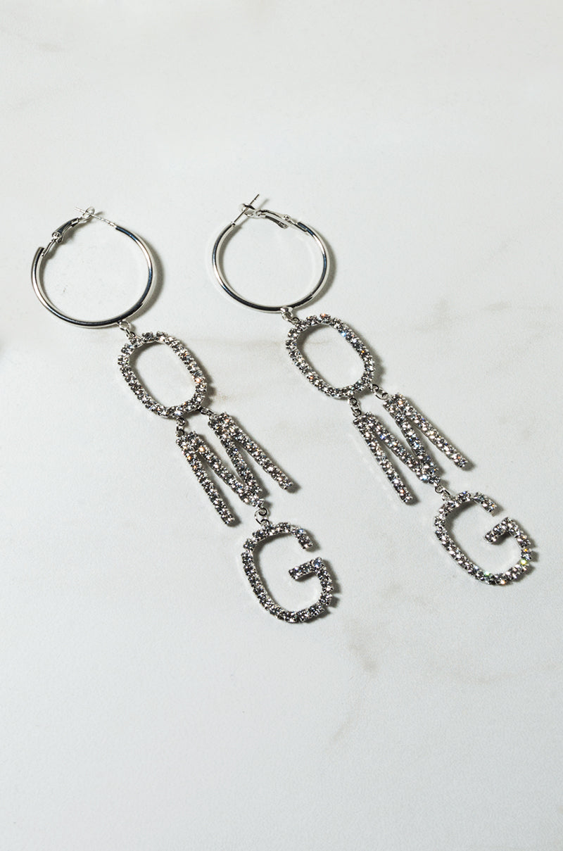 The OMG DANGLE EARRING features silver hoops adorned with dangling letters spelling "OMG," each embellished with small rhinestones and arranged vertically. The earrings are photographed lying flat on a light-colored surface.